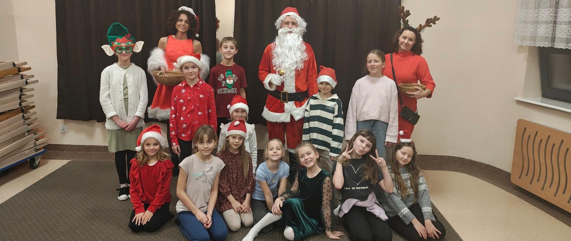 Uczniowie w czerwonych czapeczkach pozują wraz z Mikołajem oraz Śnieżynkami. Część osób siedzi, część stoi w jednej z klas szkoły muzycznej.