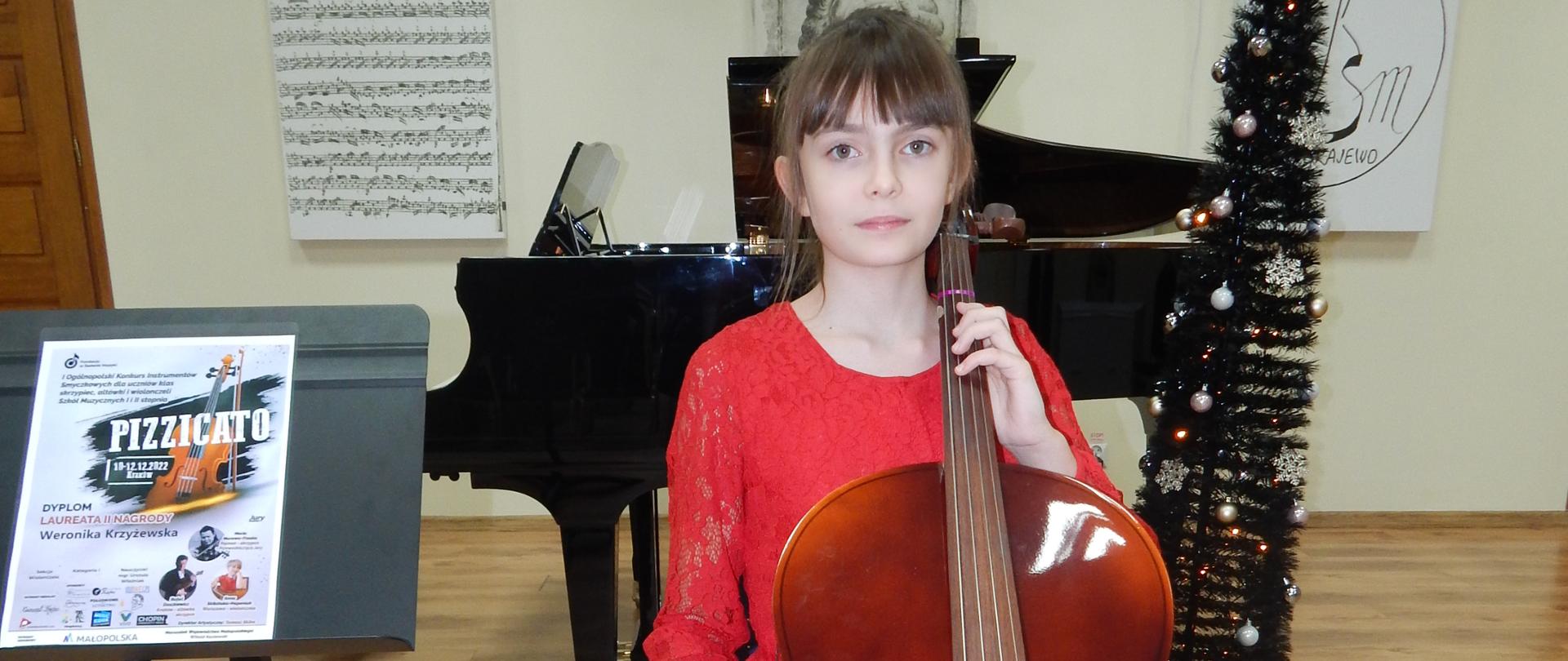 Dziewczynka ubrana w czerwoną sukienkę siedzi przy wiolonczeli , obok stoi pulpit z dyplomem za zdobycie II nagrody w kategorii I w I Ogólnopolskim Konkursie Instrumentów Smyczkowych dla uczniów klas skrzypiec, altówki i wiolonczeli
Szkół Muzycznych I i II stopnia "PIZZICATO" w Krakowie (edycja on-line). W tle fortepian i obrazy.
