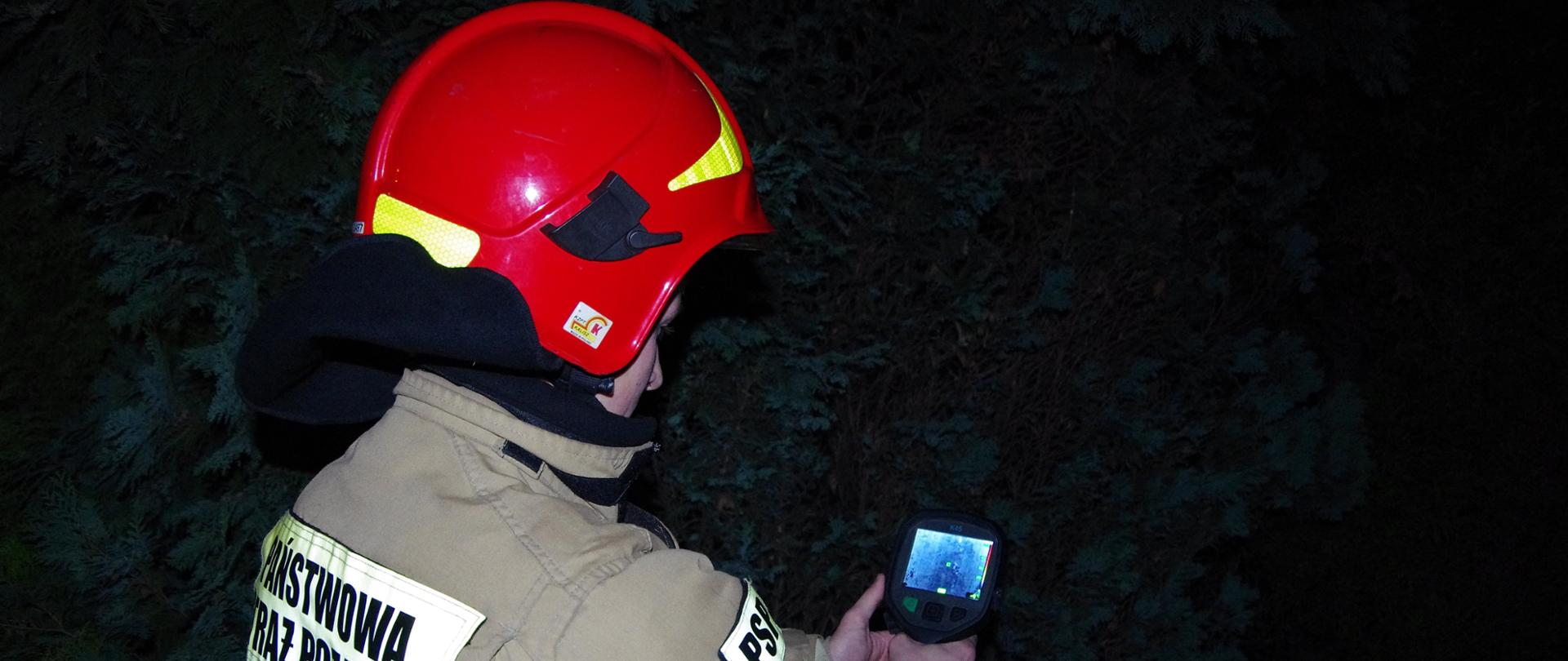 Widok z tyłu. Pora nocna - strażak w ubraniu specjalnym i czerwonym hełmie trzyma w ręku kamerę termowizyjną. 