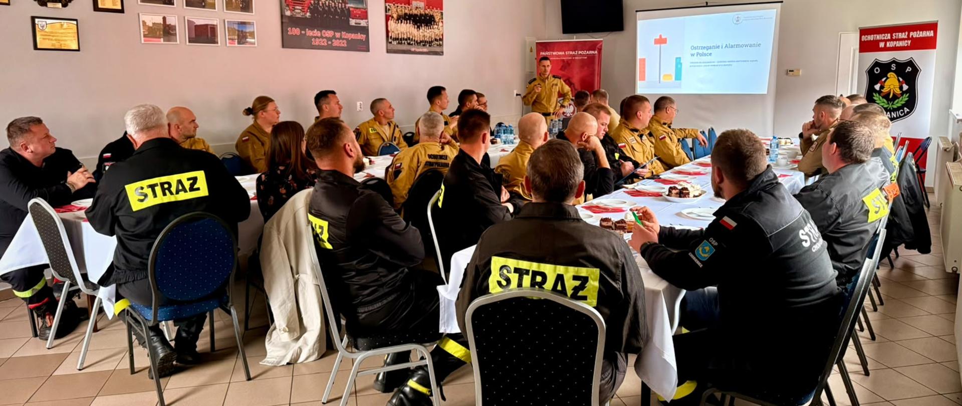 W sali spotykają się strażacy Państwowej Straży Pożarnej i Ochotniczych Straży Pożarnych. Uczestnicy siedzą przy stołach ustawionych wzdłuż pomieszczenia i słuchają prelegenta prowadzącego prezentację wyświetlaną na ekranie. Na ścianach znajdują się materiały dotyczące działalności jednostek OSP oraz oznaczenia Państwowej Straży Pożarnej. Na stołach przygotowano poczęstunek dla uczestników spotkania.