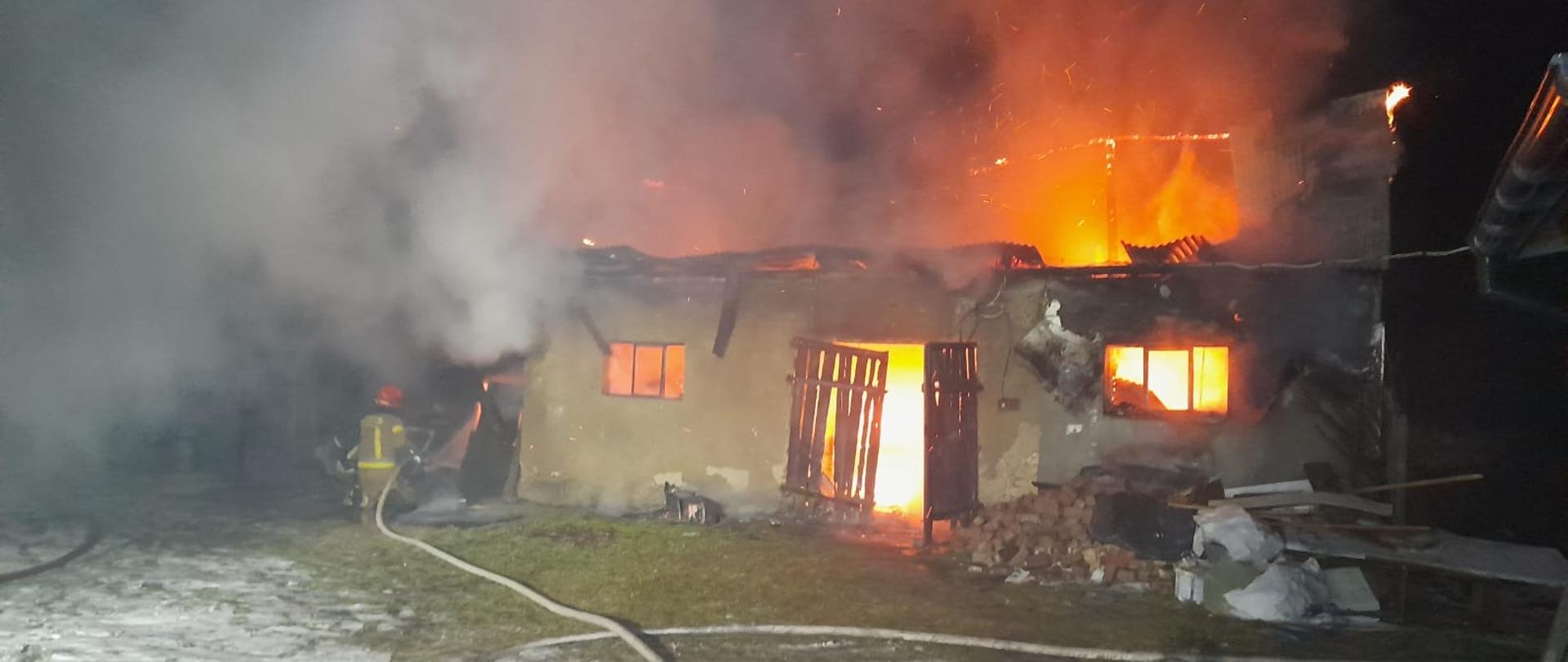 budynek płonie przez okna i drzwi widać ogień w środku dachu już prawie nie ma górą wychodzi ogień naokoło dużo dymu przed budynkiem jest strażak i gasi pożar na podwórku widać rozwinięte węże z wodą 