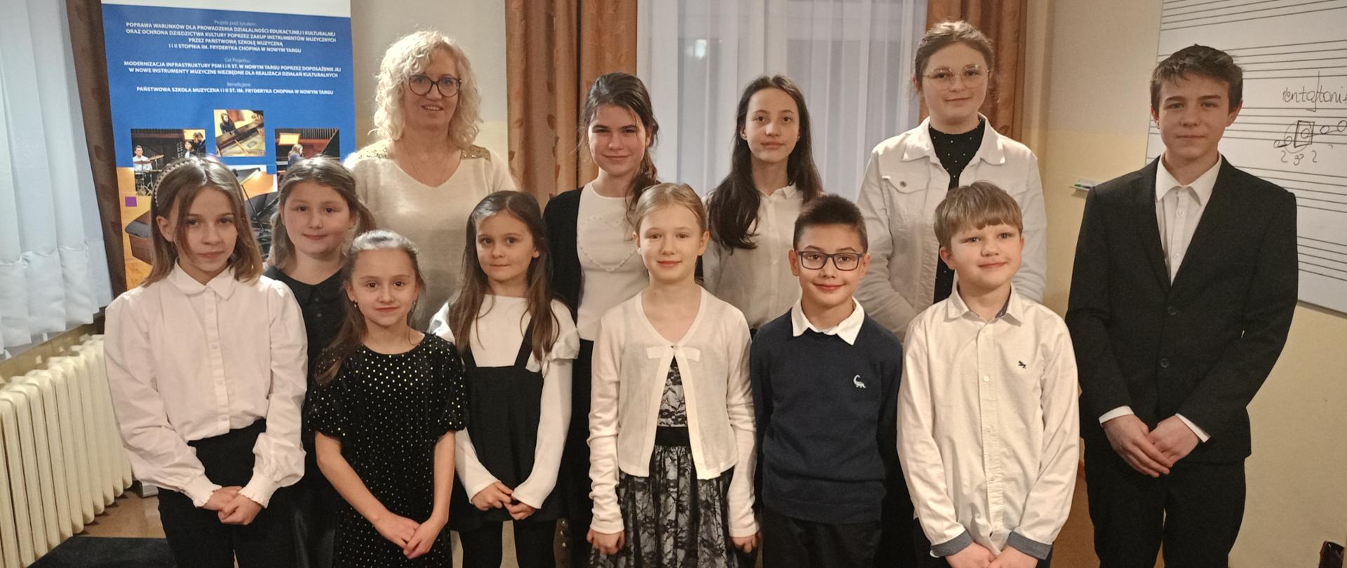 Uczniowie wraz z nauczycielką stoją w dwóch rzędach. Z lewej strony okno przysłonięte firanką i zasłonami pod nim kaloryfer a obok podnóżek fortepianowy. Z prawej strony biała tablica z pięciolinią. Za nimi okno przysłonięte firanka oraz zasłonami i baner unijny. Z sufitu zwisają dwie zapalone lampy.