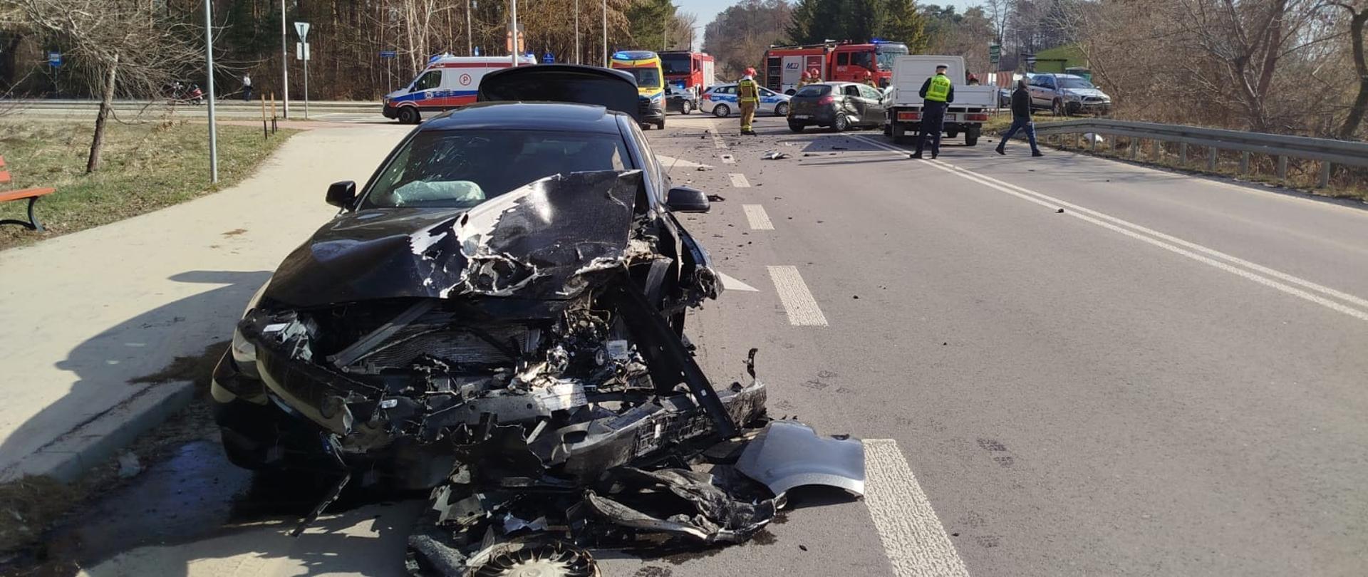 Na zdjęciu na pierwszym planie widoczny jest samochód osobowy uczestniczączy w zdarzeniu. Samochód ma ruszkodzony przód. Na drugim planie widoczne są samochody ratowniczo-gaśnicze, karetki pogotowia, radiowóz policyjny oraz funkcjonariusze służb ratowniczych.