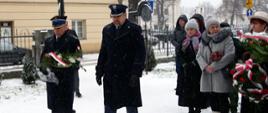 Funkcjonariusz straży pożarnej oraz straży więziennej ubrani w zimowe mundury galowe podchodzą do pomnika z wiązanka kwiatów, obok po prawo stoi sześć osób, jedna trzyma kazankę kwiatów, pasa śnieg