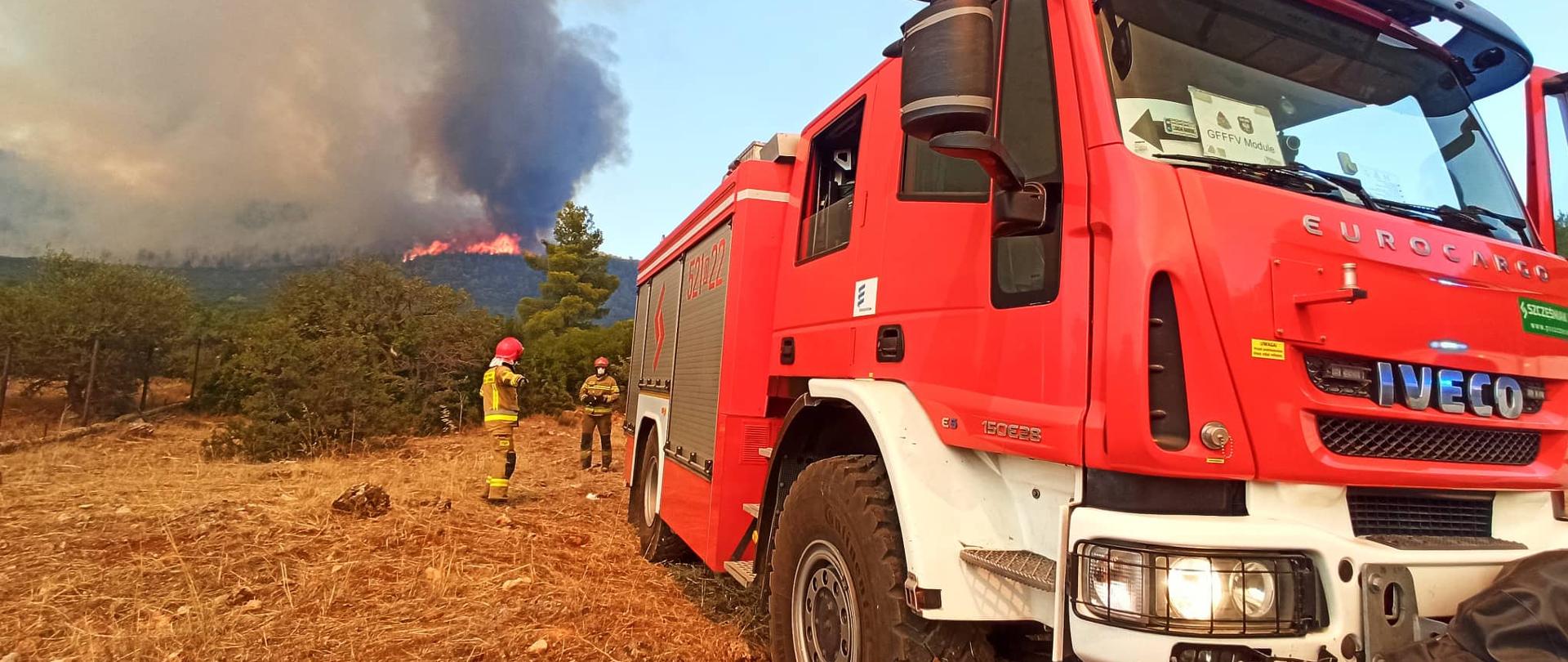 na zdjęciu widać samochód pożarniczy oraz dwóch strażaków. W tle płonące lasy w Grecji