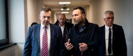 Minister Wieczorek idzie korytarzem i rozmawia z idącym obok mężczyzną w czarnym ubraniu, za nimi grupa osób.