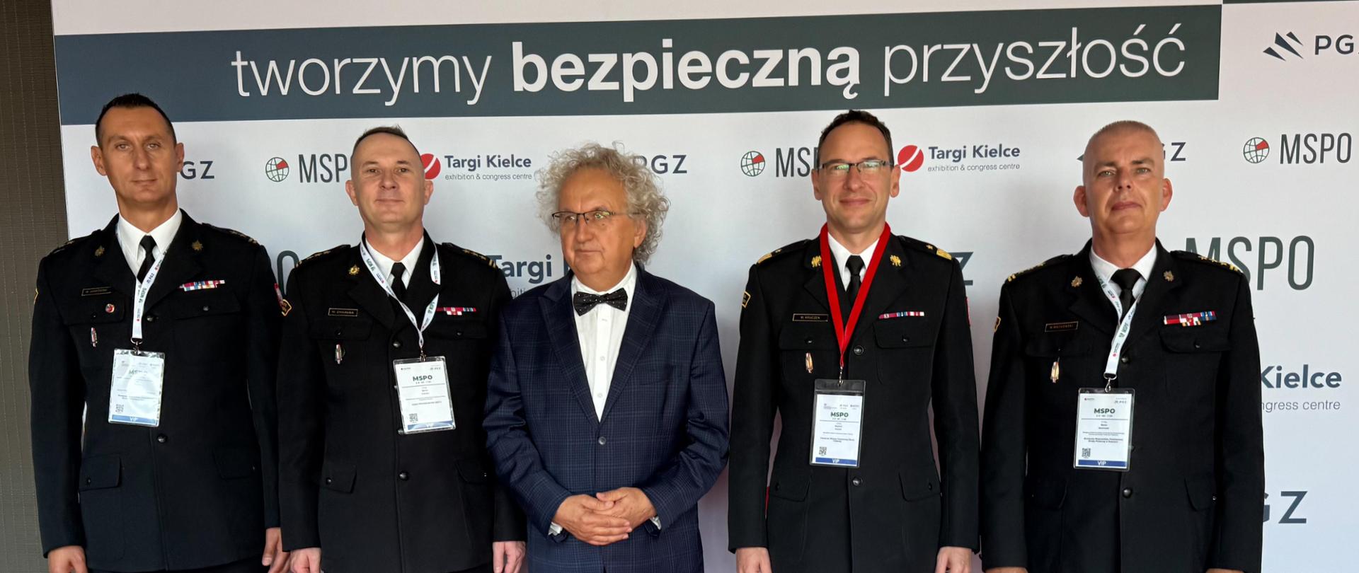 Grupa pięciu mężczyzn pozuje na czerwonym dywanie przed ścianką z logotypami targów i partnerów wydarzenia. Czterech z nich to strażacy Państwowej Straży Pożarnej w wyjściowych mundurach, a w środku stoi mężczyzna w garniturze.