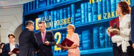 Na tle wielkiego ekranu z napisem Gala Nauki Polskiej stoi minister Wieczorek który podaje rękę mężczyźnie w czarnym garniturze, wiceminister Mrówczyńska trzymająca czerwoną teczkę i kilka osób.