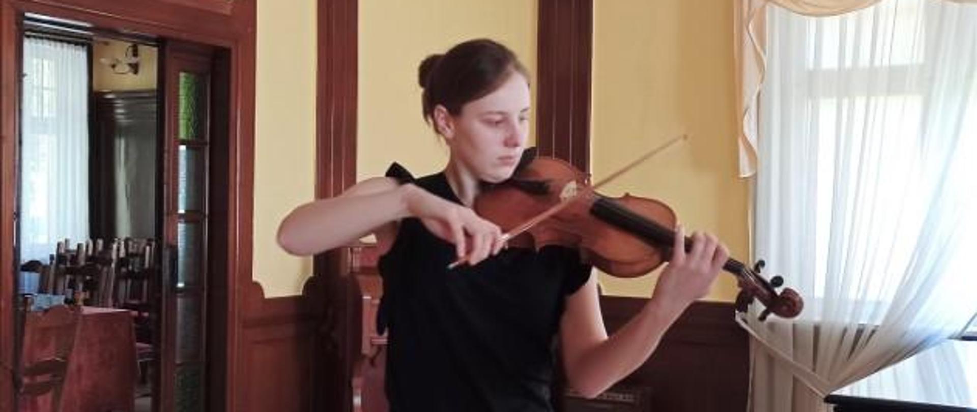 Zdjęcie kolorowe. Uczennica w czarnej bluzce i białych spodniach gra na skrzypcach. Z prawej strony fortepian i stołek do fortepianu. W tle fantazyjnie upięte zasłony.
