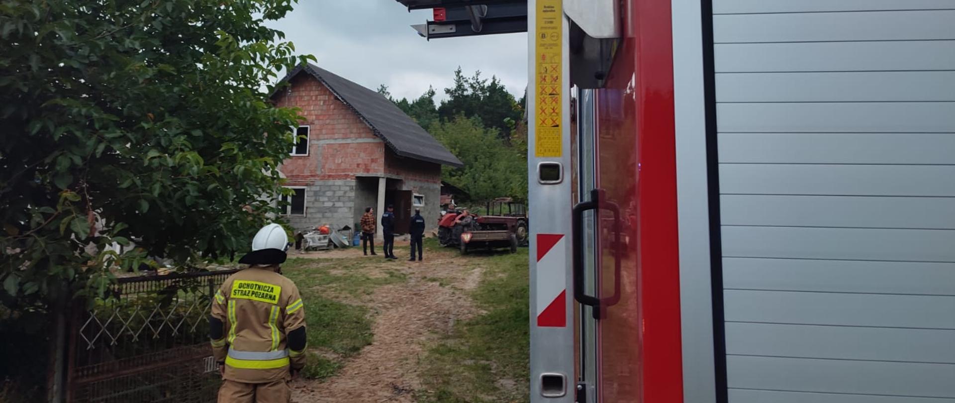 Na podwórku w oddali widać dom piętrowy z szaro-czerwonej cegły i dwuspadowym dachem pokrytym czarną papą. Za domem znajdują się drzewa. Obok domu stoi traktor oraz dwóch policjantów i osoba cywilna. Przy bramie wjazdowej na podwórko stoi strażak w piaskowym stroju strażackim i białym hełmem na głowie. Po lewej stronie od strażaka znajduje się drzewo.