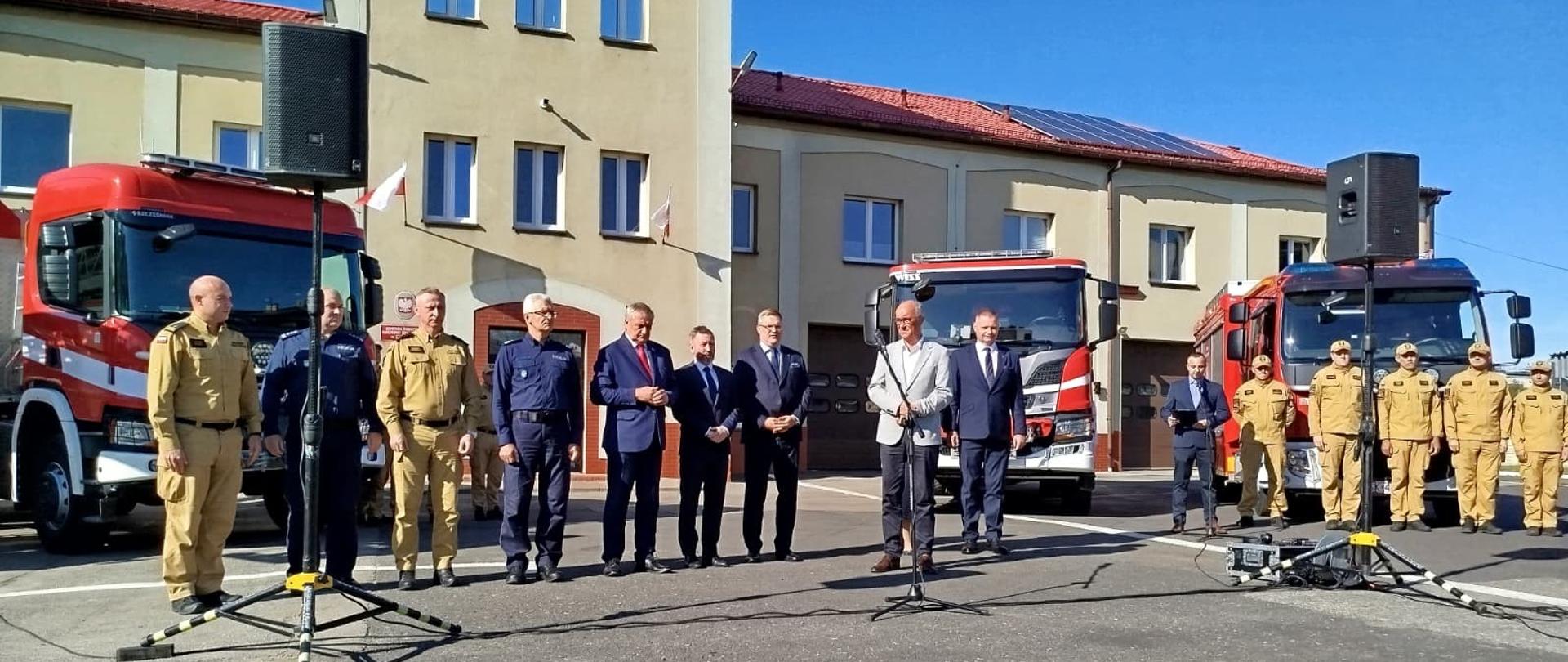 Na zdjęciu widoczne jest przygotowanie do briefingu prasowego przed budynkiem Komendy Powiatowej Państwowej Straży Pożarnej w Będzinie. W tle stoi budynek strażnicy z charakterystyczną wieżą i napisem „STRAŻ POŻARNA” oraz zaparkowane wozy strażackie. Na pierwszym planie ustawieni są funkcjonariusze straży pożarnej i policji w mundurach, a obok nich kilku przedstawicieli władz w garniturach. Po prawej stronie stoi szereg strażaków w piaskowych mundurach służbowych. Przed uczestnikami ustawione są mikrofony i głośniki. 