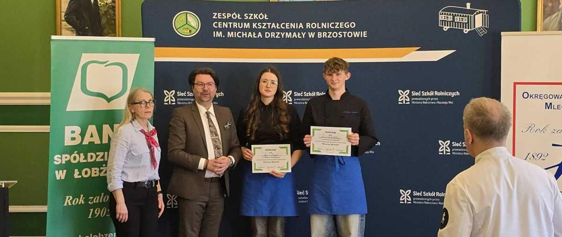 Na zdjęciu uwieczniono uroczystość wręczenia dyplomów, która odbywa się w eleganckim, klasycznym wnętrzu o zielonych ścianach, zdobionych obrazami w złotych ramach.
Na środku, przed ciemnoniebieską ścianką wystawową z logotypami szkół rolniczych, stoi grupa pięciu osób:
Po lewej stronie stoi kobieta w okularach, ubrana w jasną koszulę i ciemne spodnie.
Obok niej znajduje się mężczyzna w eleganckim brązowym garniturze i okularach, który trzyma ręce splecione przed sobą.
W centrum stoi dwoje młodych ludzi – dziewczyna i chłopak. Oboje mają na sobie ciemne bluzy i charakterystyczne, niebieskie fartuchy z logo. Z uśmiechem prezentują otrzymane dyplomy.
Z tyłu, po prawej stronie, stoi mężczyzna w czarnej koszulce, częściowo zasłonięty przez młodszego kolegę.
Na pierwszym planie, tyłem do obiektywu, widoczna jest sylwetka fotografa w białej koszuli oraz głowy innych uczestników wydarzenia.
U stóp nagrodzonych osób leżą białe torby upominkowe z logotypami. Po lewej stronie zdjęcia stoi wysoki, zielono-biały baner reklamowy ""Banku Spółdzielczego w Łobżenicy"". Nad całą sceną, na ścianie, wiszą ozdobne kinkiety i godło Polski. Atmosfera jest oficjalna, a zarazem pełna dumy z sukcesów młodych ludzi.