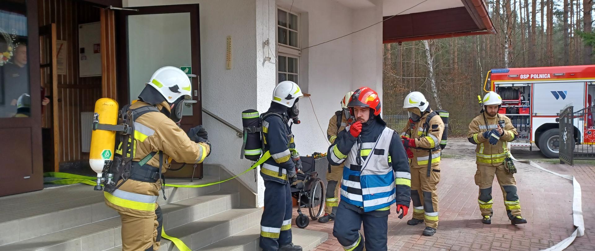 Na pierwszym planie widać strażaków w sprzęcie ochrony układu oddechowego przygotowaną do ponownego wejścia do budynku objętego pożarem oraz kierującego działaniem ratowniczym. W tle stoi samochód pożarniczy.