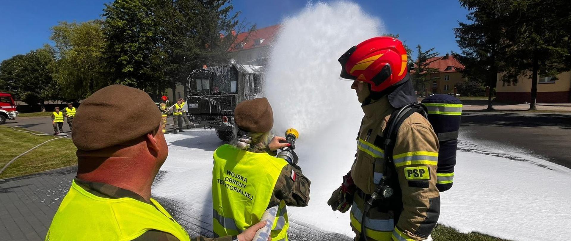Strażak w mundurze bojowym i czerwonym hełmie z prawej. Z lewej żołnierze WOT. Leja wodę z węży strażackich.