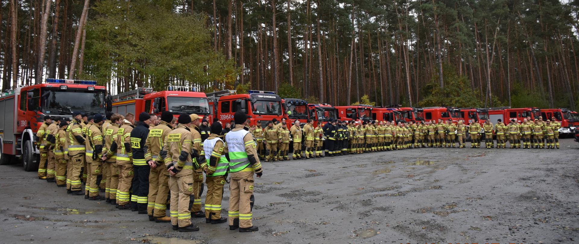 Na zdjęciu widoczne siły Wojewódzkiego Odwodu Operacyjnego Państwowej Straży Pożarnej województwa opolskiego - Kompania Gaśnicza „Opole”, Kompania Gaśnicza „Namysłów”. Grupa strażaków w ubraniach specjalnych stoi w dwuszeregu w kształcie podkowy w punkcie przyjęcia sił i środków otoczonym przez las sosnowy. W tle zaparkowanych jest kilkanaście czerwonych wozów strażackich różnych jednostek. Trwa otwarcie manewrów.