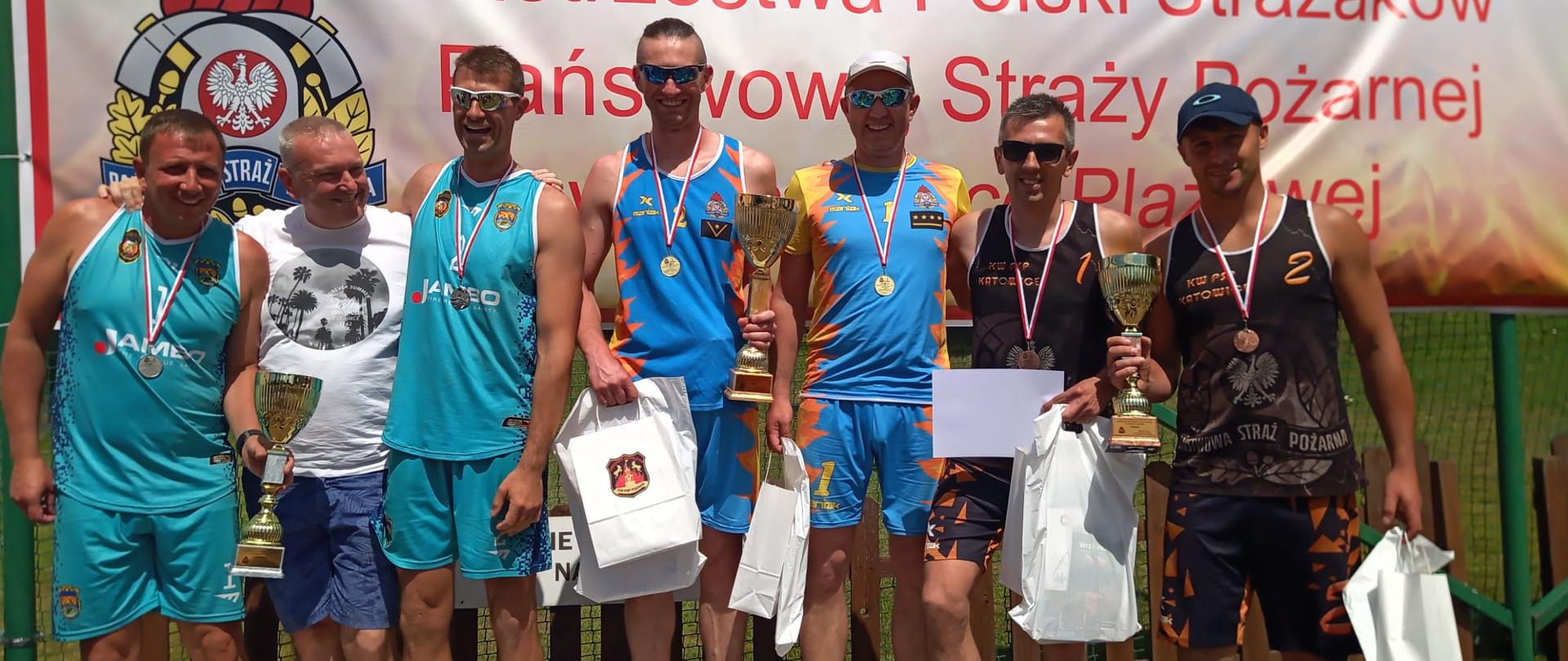Drużyny, które zajęły trzy pierwsze miejsca stoją na podium. Strażacy w ubraniach sportowych z medalami na szyi oraz pucharami w ręce. Z tyłu banner z napisem Mistrzostwa Polski Strażaków Państwowej Straży Pożarnej w siatkówce plażowej.