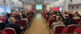 Zdjęcie przedstawia szeroki widok na salę konferencyjną podczas trwania Seminarium Bezpieczeństwa Pożarowego Budynków, ukazując widownię od tyłu.
Uczestnicy (Publiczność): Liczni uczestnicy seminarium siedzą w rzędach na czerwonych krzesłach, skierowani w stronę sceny. Widać, że na widowni dominują dorośli, w tym wiele kobiet i mężczyzn w różnym wieku, co jest zgodne z informacją o udziale zarządców, administratorów budynków i mieszkańców.
Scena i Prelegent: W centralnej części tła znajduje się scena. Na dużym ekranie widać logo Państwowej Straży Pożarnej i symbol kampanii "Czujka na straży Twojego bezpieczeństwa!". Na scenie, po prawej stronie, widać prelegenta (prawdopodobnie w mundurze PSP lub stroju cywilnym) stojącego przy mównicy i przemawiającego do zebranych.
Wnętrze: Sala ma jasny sufit z nowoczesnym oświetleniem punktowym i jasną, drewnianą podłogę. Po lewej stronie, z tyłu sali, stoją dwie osoby (jedna w mundurze) obserwujące wydarzenie.
Zdjęcie, z centralnie poprowadzonymi rzędami krzeseł, podkreśla formalną i edukacyjną atmosferę spotkania, dokumentując duże zainteresowanie tematyką bezpieczeństwa pożarowego.