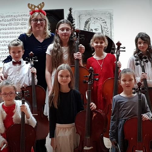 Uczniowie stoją , każdy z wiolonczelą , za nimi nauczycielka, w tle widać obrazy