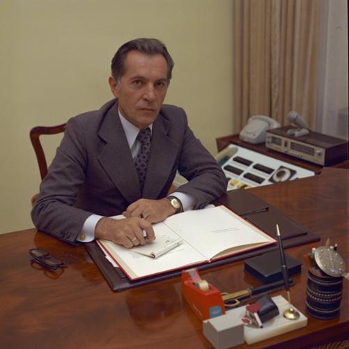 Emil Wojtaszek - kolorowy portret mężczyzny siedzącego przy drewnianym biurku, ciemne włosy, grafitowa marynarka, ciemny krawat, biała koszula. Na biurku przybory do pisania, kalendarz i okulary.