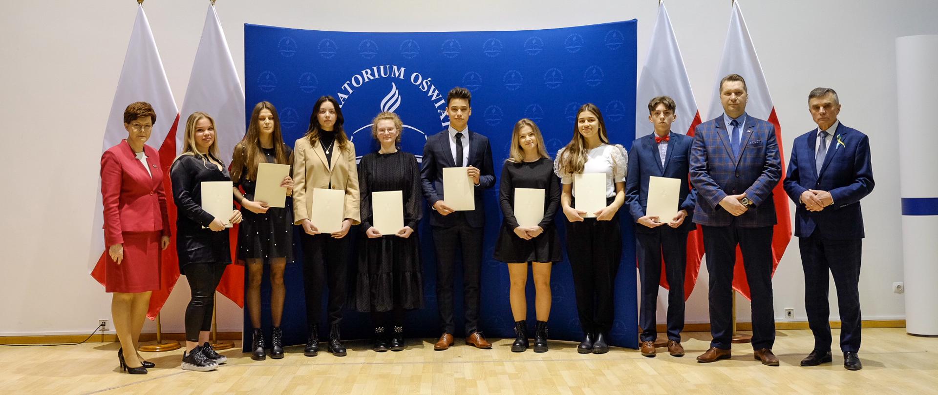Zdjęcie zbiorowe - stypendyści stoją pod ścianą, trzymają w rękach dyplomy.