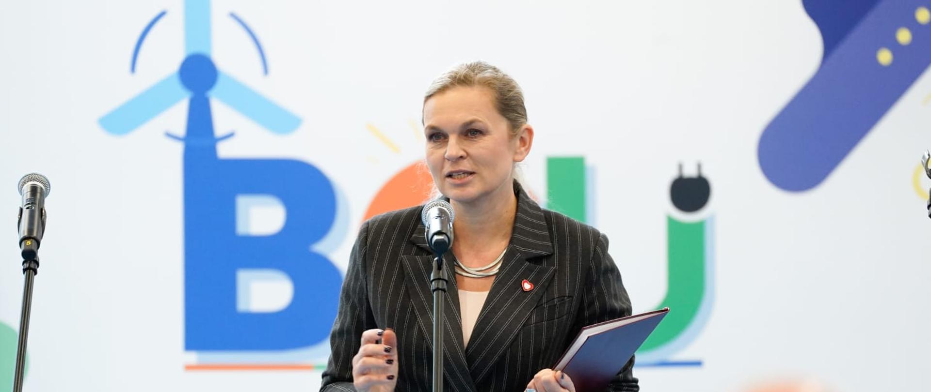 Minister Edukacji Barbara Nowacka w garniturze w prążki, trzymająca czerwoną teczkę, przemawia do mikrofonu na tle dużego, kolorowego banera promującego szkolnictwo branżowe i odnawialne źródła energii.