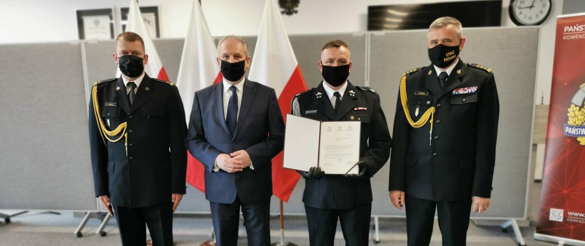 Na zdjęciu znajduje się 3 strażaków umundurowanych w mundury galowe oraz mężczyzna w granatowym garniturze. Strażak ubrany w mundur galowy OSP trzyma w rękach przed sobą dokument z promesą. Za mężczyznami ustawione są trzy flagi Polski.