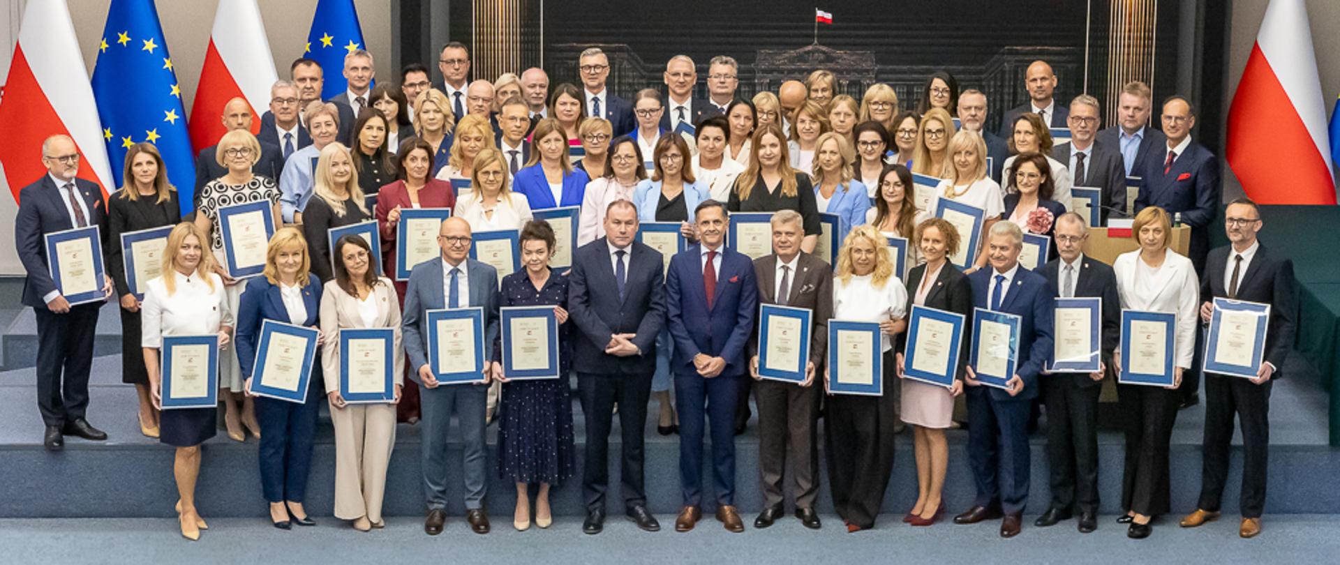 Laureaci stoją z dyplomami na tle ścianki, w tle flagi
