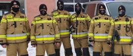 6 strażaków PSP ubranych w nowy wzór ubrania specjalnego pozuje do zdjęcia na dworze, na ziemi leży śnieg, za strażakami stoi samochód , w tle budynek strażnicy