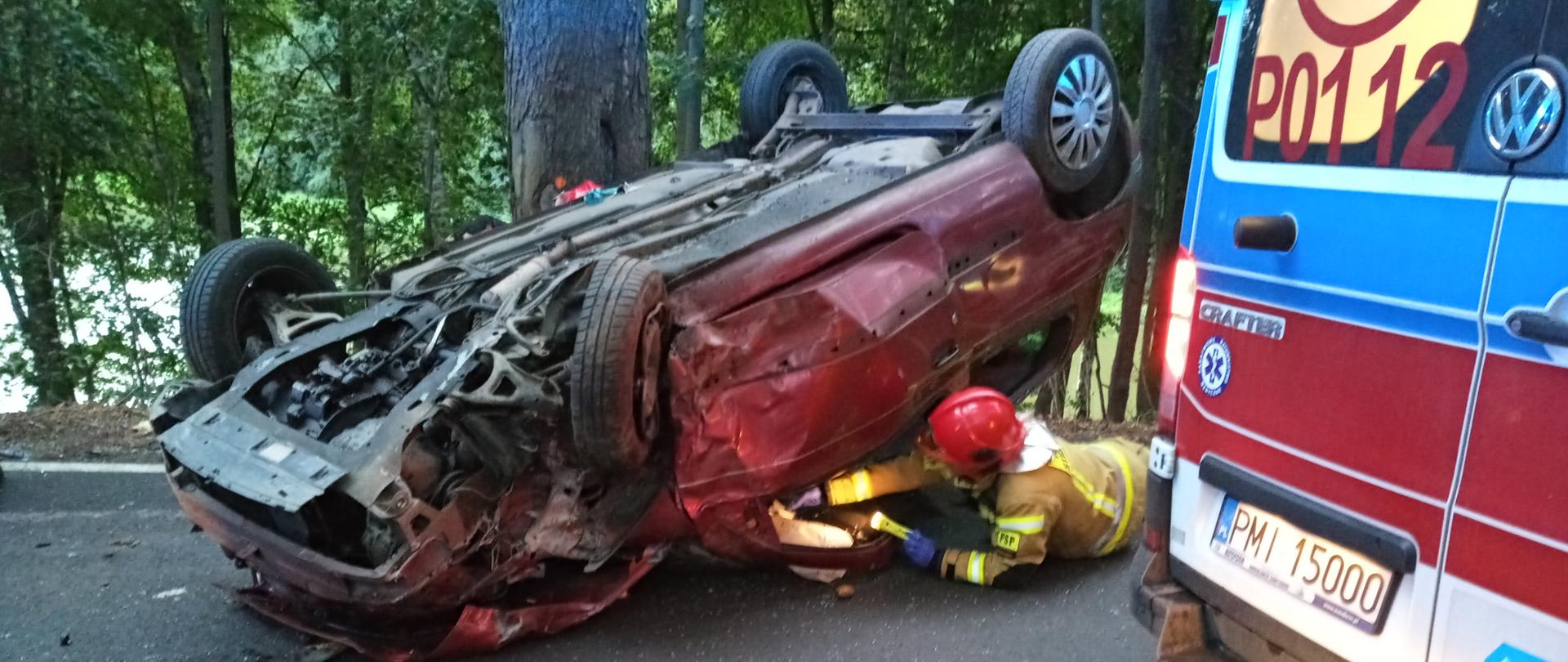 Samochód osobowy leży na dachu , strażak kładąc się na ziemi sprawdza wnętrze pojazdu.