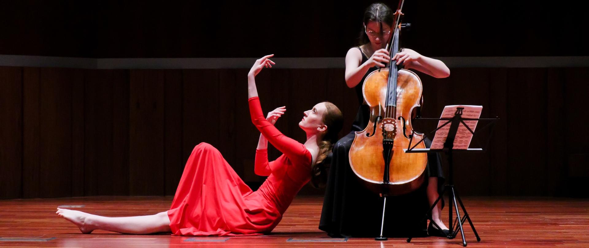 Na scenie dwie młode kobiety w czeronej i czarnej sukience - kobieta w czarnej gra na wiolonczeli podczas gdy kobieta w czerwonej sukience przybiera pozę siedzącą - prawie opartą o wiolonczelistkę