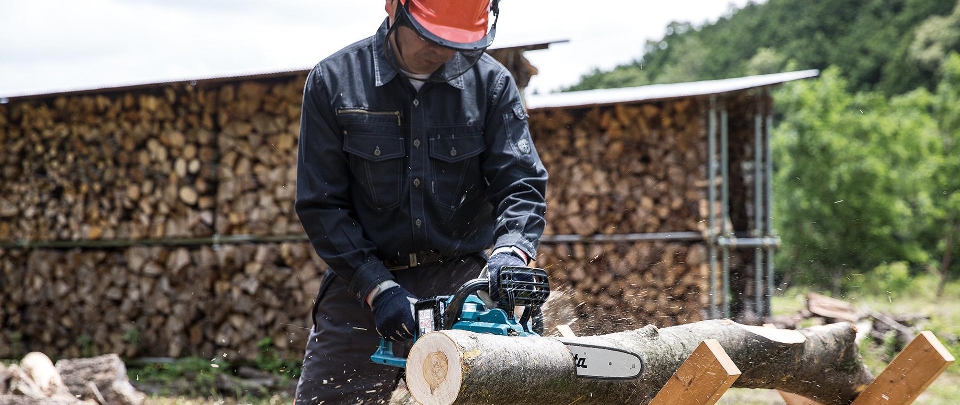 Mężczyzna w ochronnikach słuchu i kasku w pozycji stojącej na koziołku drewnianym piłuje konar drewna przy użyciu piły spalinowej