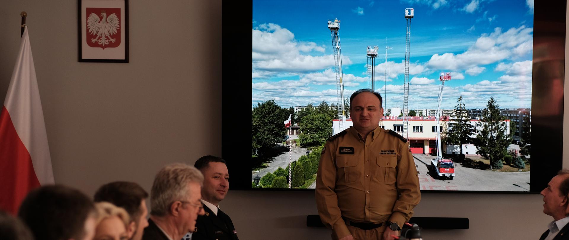 Komendant powiatowy przemawia do gości zgromadzonych na spotkaniu. w tle na ścianie wyświetlacz z obrazem podnośników strażackich oraz godłem Polski.