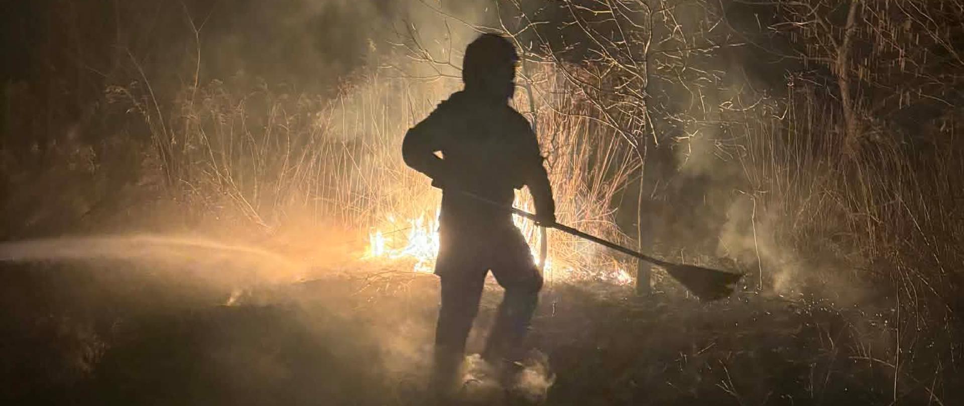 Sylwetka osoby gaszącej ogień nocą; oświetlona płomieniami i otoczona dymem.