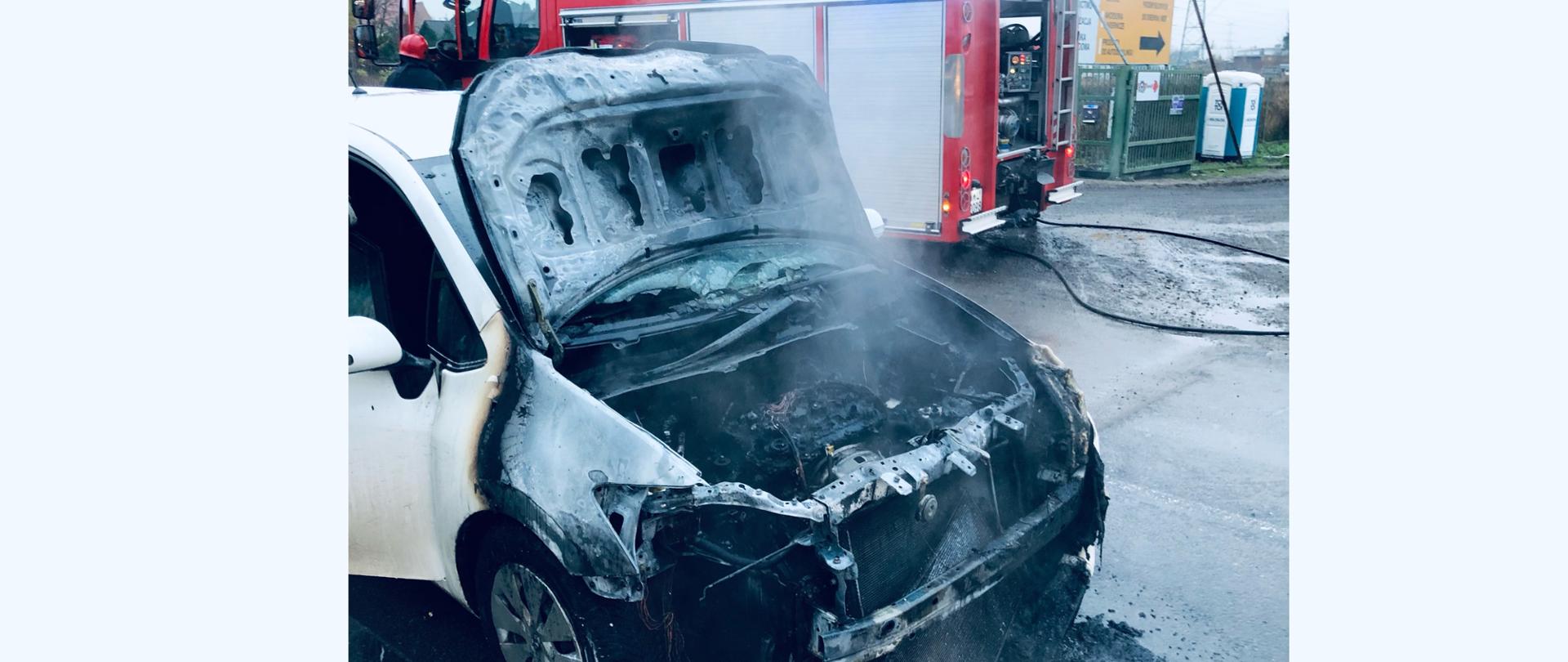Zdjęcie zrobione na tle samochodu strażackiego. Na zdjęciu spalone auto osobowe, w którym spaleniu uległa komora silnika