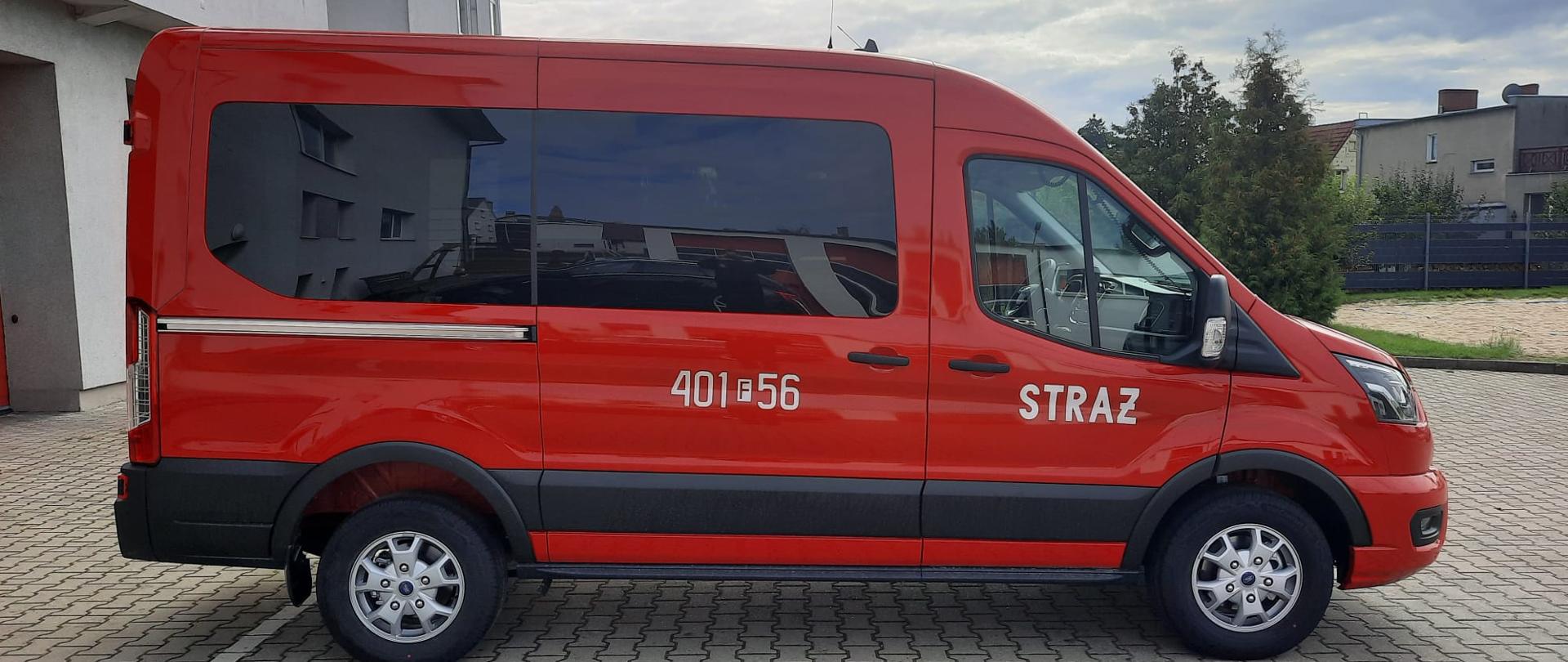 Fotografia przedstawia Mikrobus Ford Transit w kolorze czerwonym stojący na placu. Dalej za pojazdem po lewej stronie ujęcia widoczny budynek Jednostki Ratowniczo-Gaśniczej PSP w Strzelcach Krajeńskich, w tle zachmurzone niebo. Ujęcie prawego boku pojazdu. 