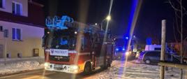 Na zdjęciu widać długi rząd pojazdów strażackich biorących udział w nocnej akcji. Śnieg i odbicia światła na drodze podkreślają trudne warunki, w jakich pracują ratownicy. Widoczne są zaparkowane samochody oraz hydrant w pobliżu, co może sugerować pobieranie wody do działań gaśniczych. W tle widać budynki mieszkalne, co może świadczyć o zagrożeniu dla mieszkańców. Światła sygnalizacyjne dodają dynamiki zdjęciu.
