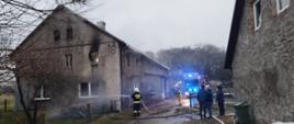 Dzień . Z lewej strony budynek mieszkalno-gospodarczy. Widać duże okopcenie wokół jednego z otworów okiennych na II kondygnacji, nad dachem budynku unosi się para. Na ziemi przy ścianie leż sterty nadpalonych lub spalonych drewnianych desek i elementów wyposażenia domu. Stojący obok strażak w granatowym ubraniu specjalnym i białym hełmie podaje prąd wody na stertę spalonych elementów. W tle obok budynku stoi samochód specjalny - drabina mechaniczna. Z prawej strony zdjęcia widać część ściany budynku, obok którego stoją trzy osoby w ubraniu cywilnym. Na ziemi leży kilka rozwiniętych węży strażackich
