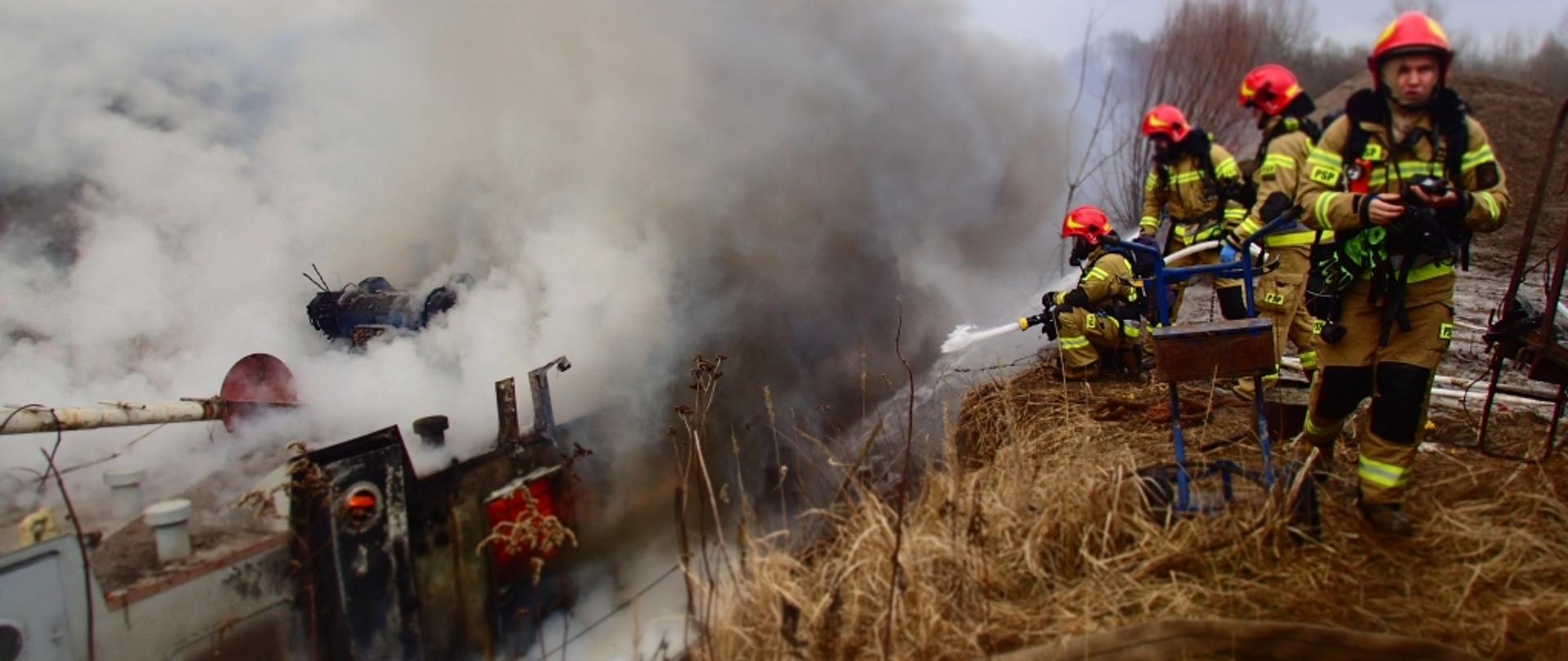 Na zdjęciu po prawej stronie widoczni są ratownicy PSP. Znajdują się oni na skarpie nad zatoką przy Wiśle. Po lewej stronie zdjęcia pali się barka zacumowana do brzegu. Wydobywa się z niej gęsty dym. Za jednym z okien barki widać płomienie