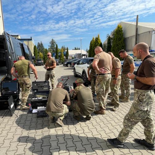 Na brukowanym parkingu obok półciężarówki grupa mężczyzn ogląda sprzęt umieszczony w kilku walizkach