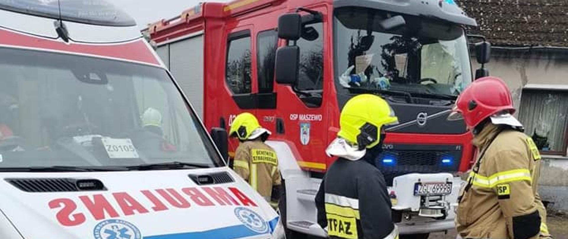Na zdjęciu widocznych dwóch strażaków w ubraniu specjalnym stojących przed karetką Państwowego Ratownictwa Medycznego oraz dwóch strażaków stojących przy wozie bojowym OSP Maszewo.
