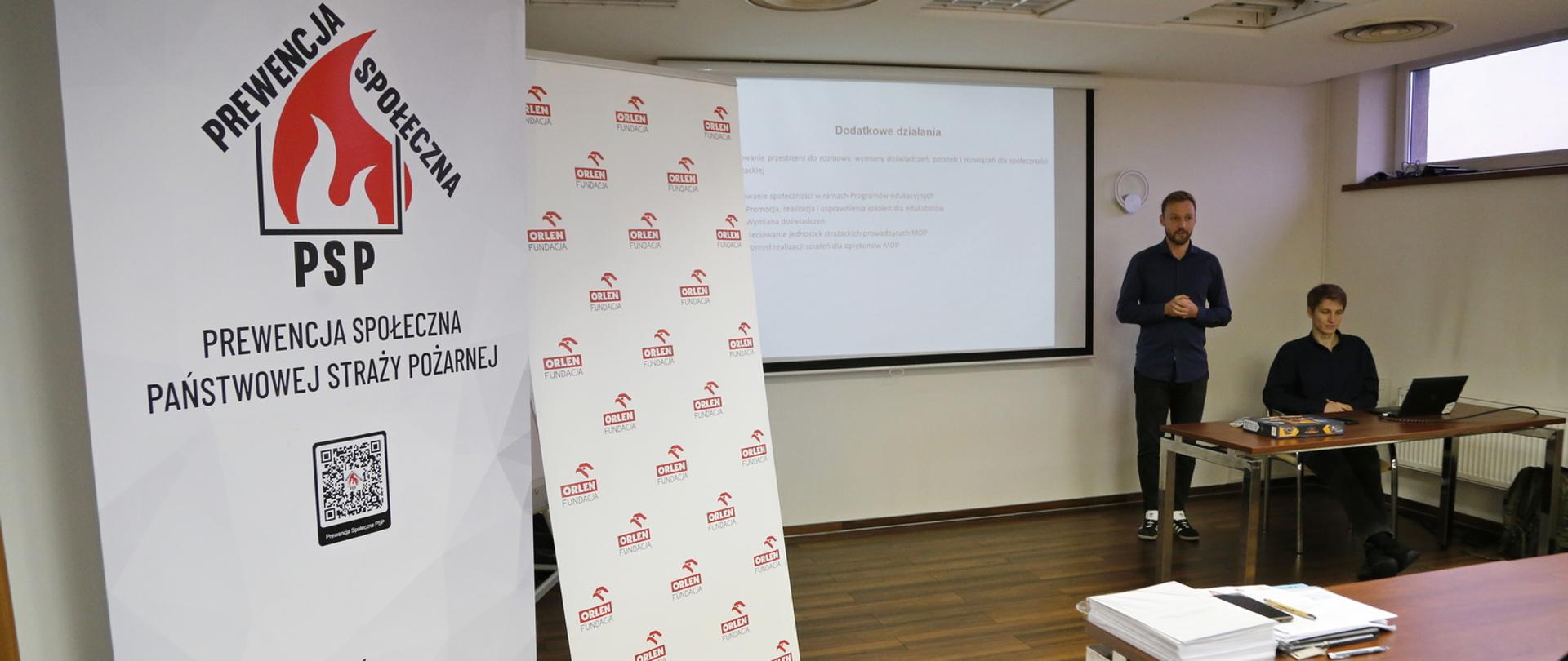 Po lewej biały baner z umieszczonym na górze logo kampanii Prewencja społeczna PSP, dalej baner z logo firmy Orlen, w głębi stół przy którym siedzi kobieta, za nią stoi mężczyzna
