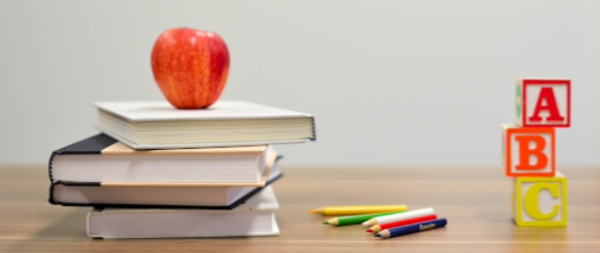 Fotografia przedstawiająca blat stołu ze stosem kolorowych książek, na których leży czerwone jabłko. Obok rozsypane kolorowe ołówki oraz wieża zbudowana z klocków zawierających symbole liter A, B, C