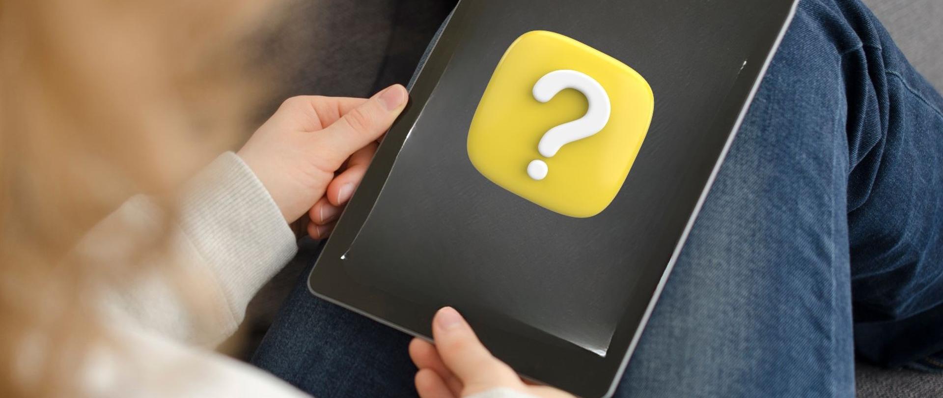 Zdjęcie przedstawia kobietę ubraną w ciemne jeansowe spodnie oraz beżową bluzę która trzyma w rękach tablet na którego ekranie widać na żółtym tle biały znak zapytania. Nie widać jej twarzy. 