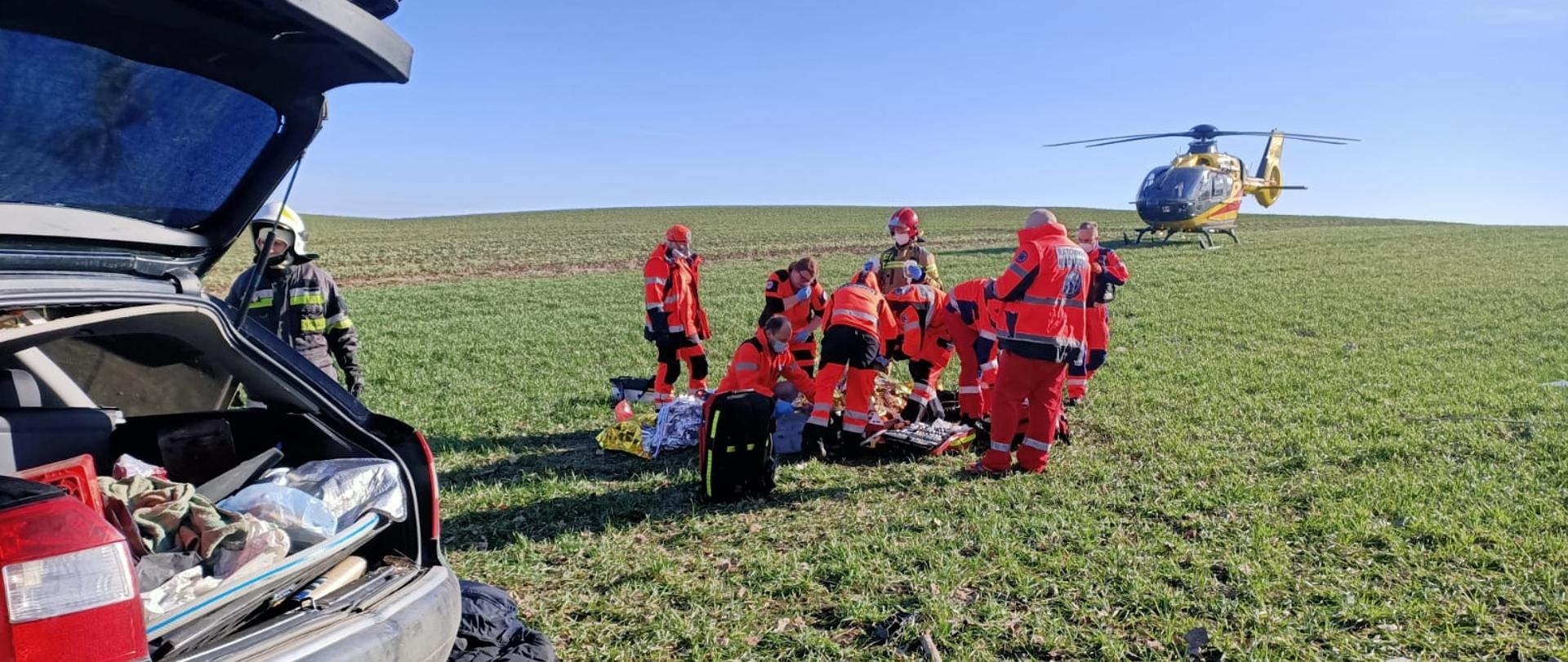 Obraz przedstawia ratowników medycznych i strażaków udzielających pomocy medycznej osobie poszkodowanej. Na pierwszym palnie uszkodzony samochód osobowy. W tle śmigłowiec Lotniczego Pogotowia Ratunkowego.