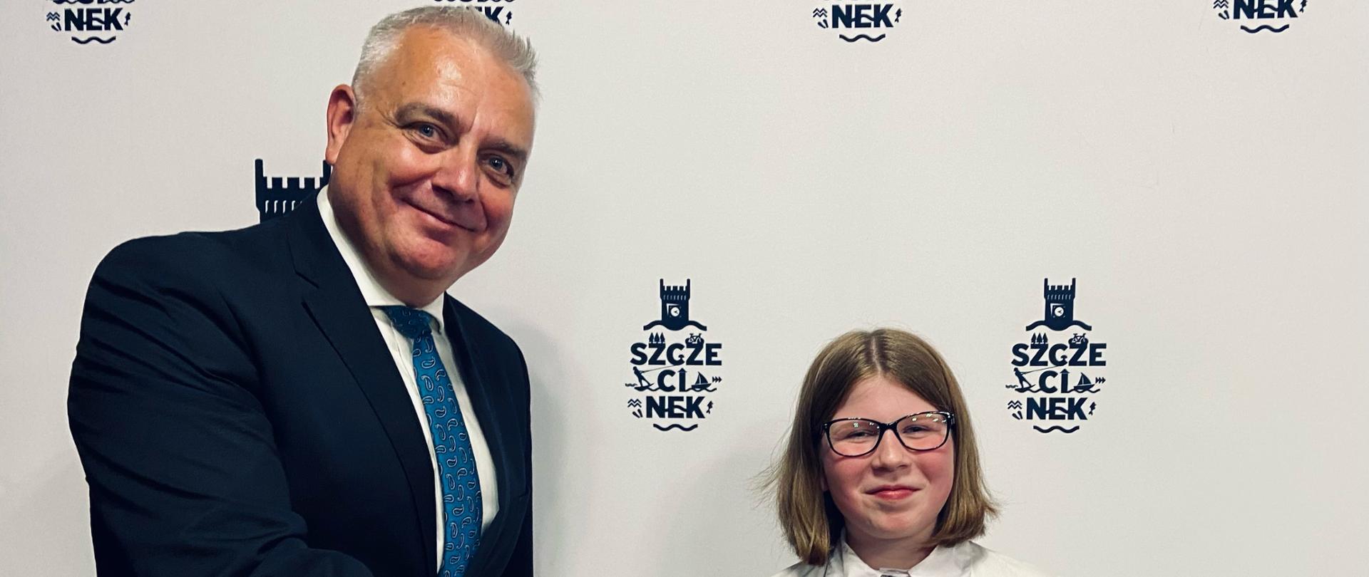 Zdjęcie przedstawia: na jasnym tle w czarne loga miasta Szczecinek. Od lewej postać mężczyzny w średnim wieku, ubranego w czarny garnitur, białą koszulę i niebieski krawat w okrągłe wzorki, który ściska dłoń 10-cio letniej dziewczyny w okularach o włosach koloru ciemny-blond. Dziewczyna trzyma w lewej dłoni ciemnozieloną teczkę z żółtym herbem miasta Szczecinek i konturami ratusza. pod spodem dziewczyna trzyma szarą, papierową torbę