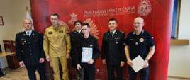 fotografia przedstawia laureata wraz z kadrą oraz egzaminatorami konkursu, w tle ścianka Państwowa Straż Pożarna, część wyposażenia Sali