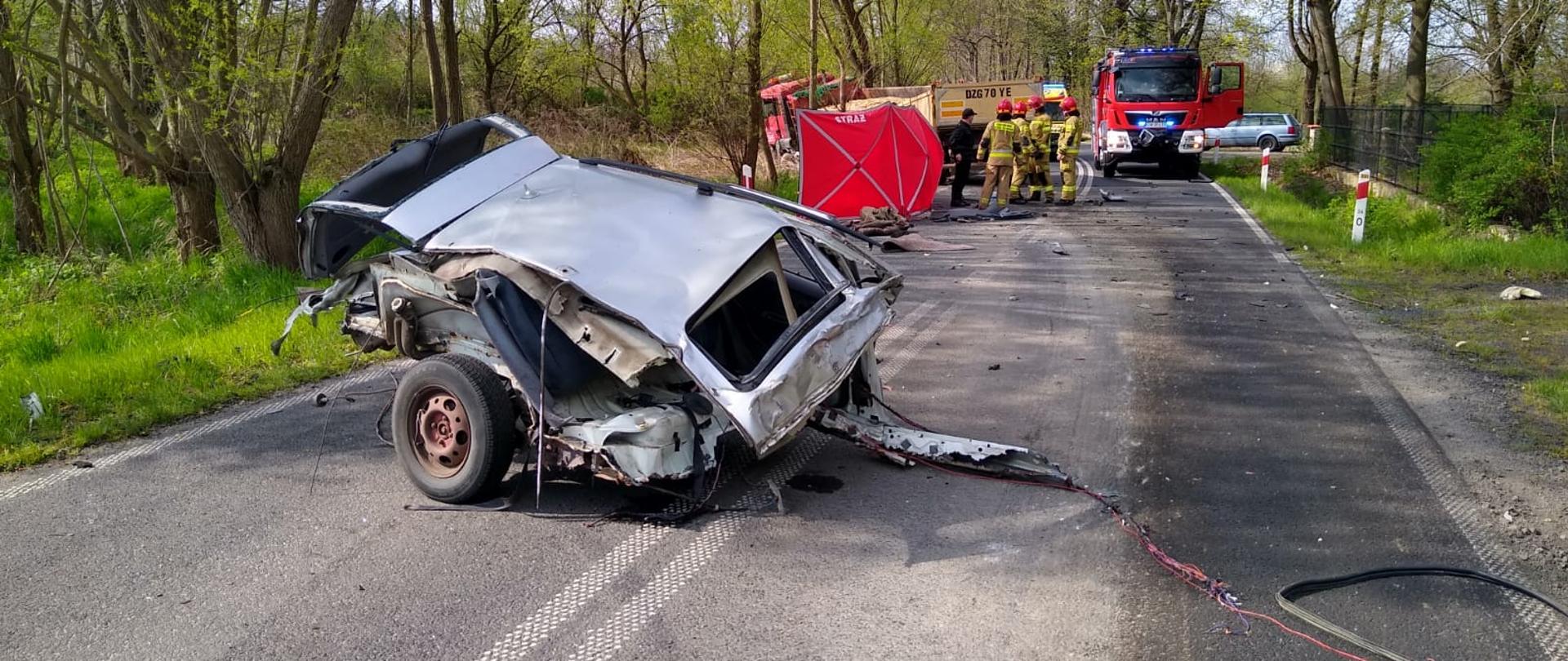 Na pierwszym planie tylna część rozbitego pojazdu osobowego, w tle grupa strażaków stoi przy czerwonym parawanie. Dalej samochód ratowniczo-gaśniczy oraz samochód ciężarowy znajdujący się poza pasem drogi