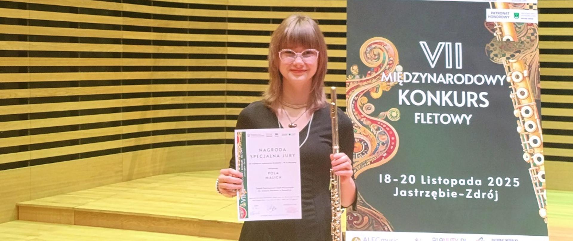 Uczennica trzyma w ręku flet poprzeczny i dyplom, na tle banera reklamowego konkursu i drewnianej ściany