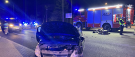 Na pierwszym planie przód uszkodzonego samochodu osobowego na jezdni, za nim pojazd gaśniczy, policjant i strażak. Pora nocna.