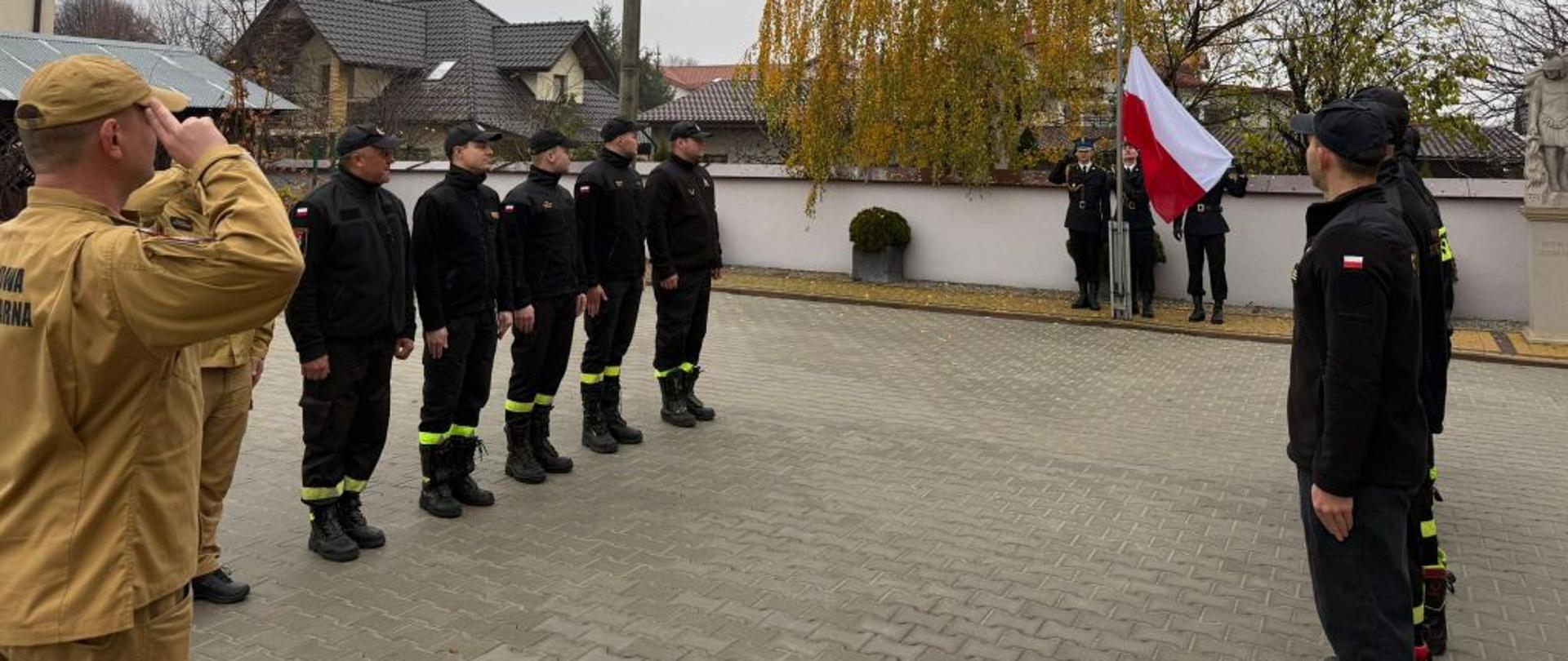 Na zdjęciu dwie zmiany służbowe podczas zmiany służby na placu przed Komendą Powiatową Państwowej Straży Pożarnej w Szydłowcu. W oddali poczet flagowy wciągający flagę państwową na maszt. 